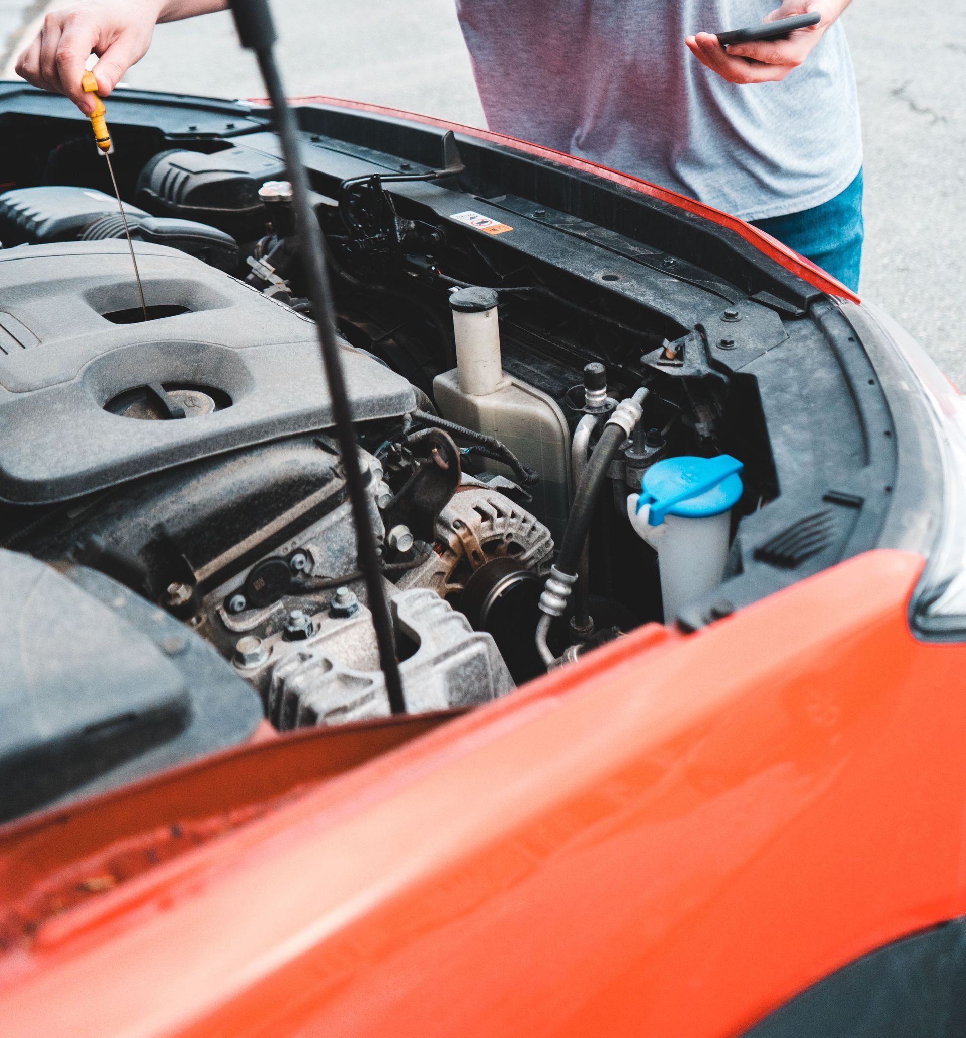 Man inspecting car engine while holding phone outdoors, checking oil levels.