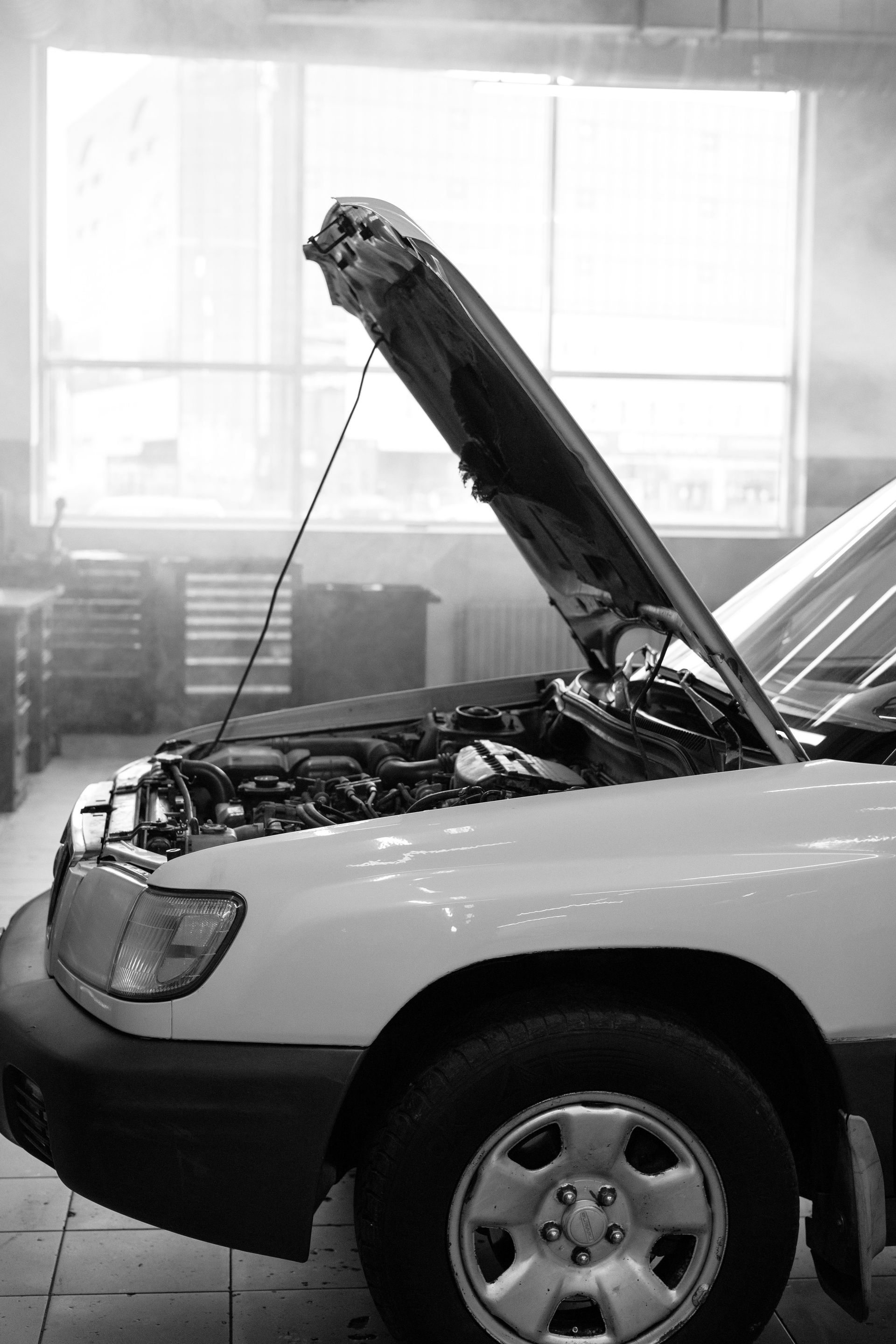 Black and white photo of a car with open hood in an auto repair shop, capturing the repair process.